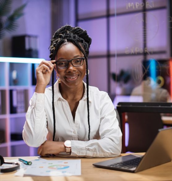 Front view portrait of young smiling experienced smart stylish african american female office manage Front view portrait of young smiling experienced smart stylish african american female office manager in white shirt sitting at table using laptop in evening office.